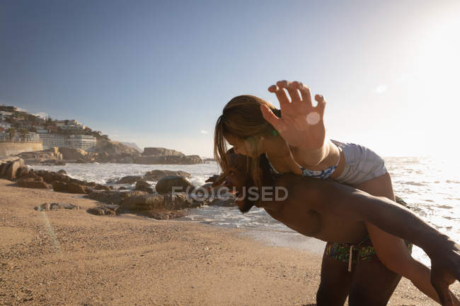 Vista lateral de un joven afroamericano llevando a una mujer a cuestas en la playa en un día soleado - foto de stock