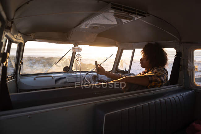 Rückansicht eines Mixed-Race-Mannes, der in einem Wohnmobil auf sein Handy schaut, gegen den Strand mit Sonnenuntergang im Hintergrund — Stockfoto