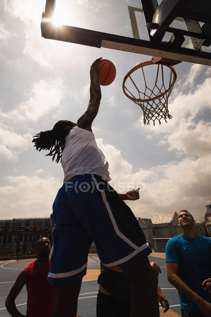 Rückansicht eines afrikanisch-amerikanischen Basketballspielers, der springt, um einen Korb zu treffen, während andere Spieler ihn auf einem Basketballfeld betrachten — Stockfoto
