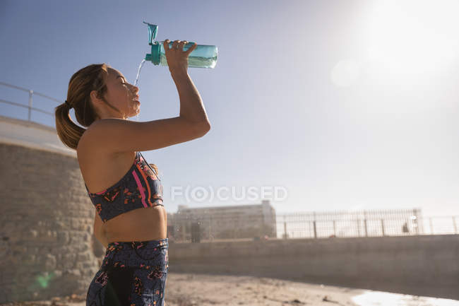 Vista lateral da mulher derramando água em seu rosto após o exercício na praia em um dia ensolarado — Fotografia de Stock