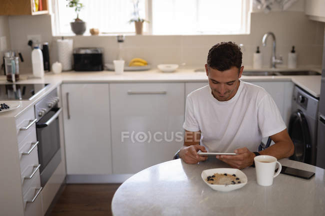 Vista frontal do jovem caucasiano usando tablet digital enquanto toma café da manhã na cozinha em casa — Fotografia de Stock