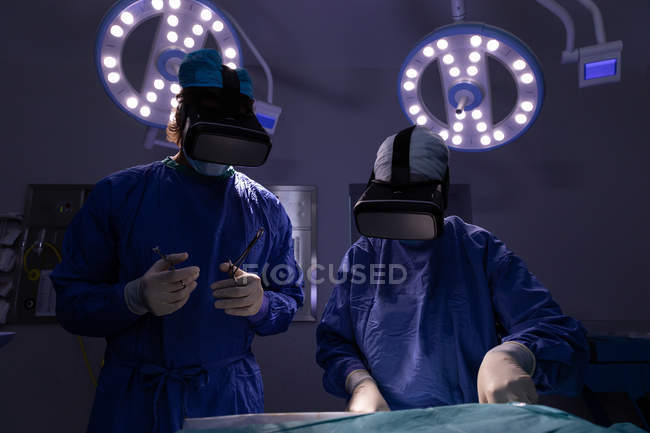 Вид спереди хирургов, использующих гарнитуру виртуальной реальности во время операции в операционной в больнице с пятнами над ними — стоковое фото