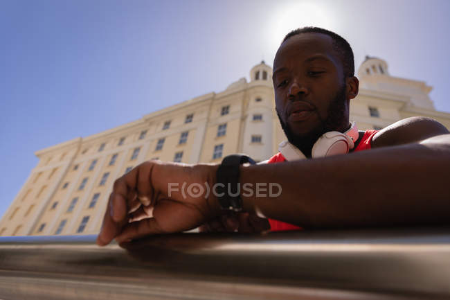 Visão de baixo ângulo do jovem afro-americano em forma de homem com fone de ouvido olhando para relógio inteligente enquanto está de pé por trilhos — Fotografia de Stock