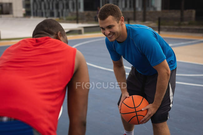 Vista lateral de jugadores de baloncesto multiétnicos que interactúan entre sí mientras juegan baloncesto en la cancha de baloncesto. Están sonriendo. - foto de stock