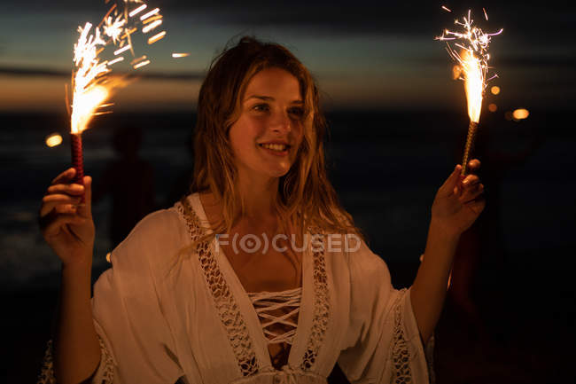 Front view of beautiful Caucasian woman standing with sparkler on beach at dusk — Stock Photo
