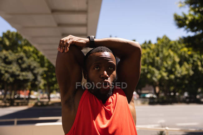 Vista frontal de un joven afroamericano en forma haciendo ejercicio de estiramiento manual en el pavimento bajo el puente en un día soleado - foto de stock
