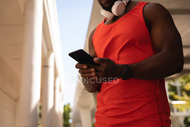 Vista lateral do jovem homem apto afro-americano com fone de ouvido usando telefone celular enquanto está de pé na rua — Fotografia de Stock