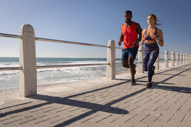Frontansicht eines jungen multiethnischen Paares, das an einem sonnigen Tag auf dem Bürgersteig in der Nähe des Promenadenstrandes läuft — Stockfoto
