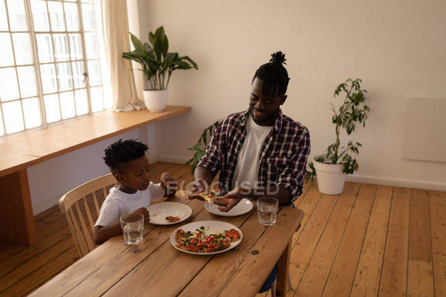 Front view of happy African-American father and son having pizza at home — Stock Photo