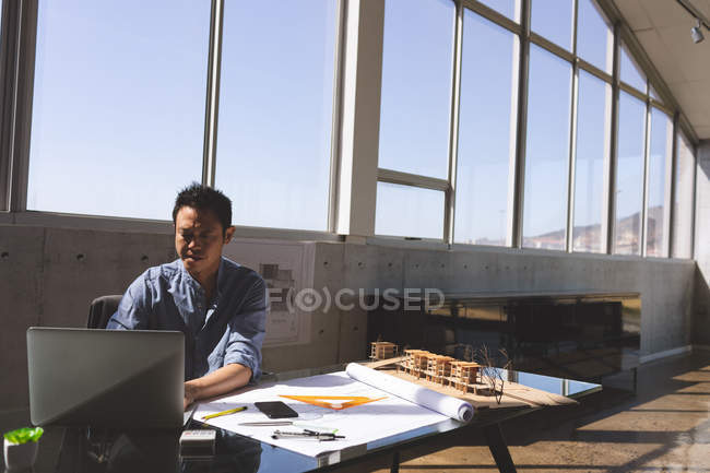 Vue de face d'un architecte masculin asiatique travaillant dur à l'aide d'un ordinateur portable tout en travaillant sur un plan avec règle triangle orange, boussole géométrique et crayons au bureau dans un bureau moderne contre le ciel bleu en arrière-plan — Photo de stock