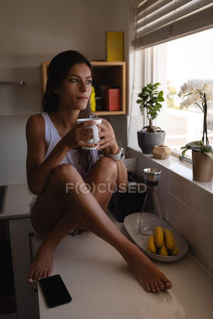 Вид спереду красива змішаної раси жінка п'є каву, сидячи на кухні вдома. Вона дивиться геть — стокове фото