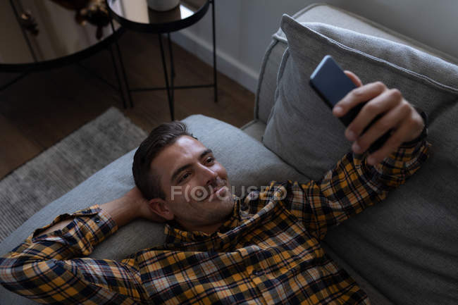 Vista de ángulo alto del hombre caucásico utilizando el teléfono móvil mientras se apoya en el sofá en casa - foto de stock