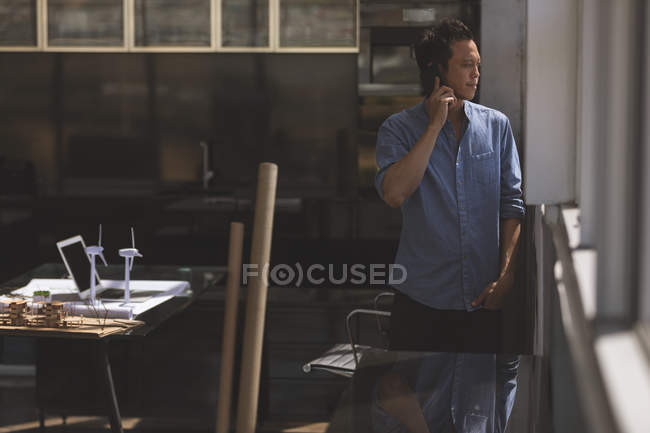 Vorderansicht eines asiatischen männlichen Architekten, der im Stehen mit dem Handy telefoniert und in einem modernen Büro auf das Fenster blickt — Stockfoto