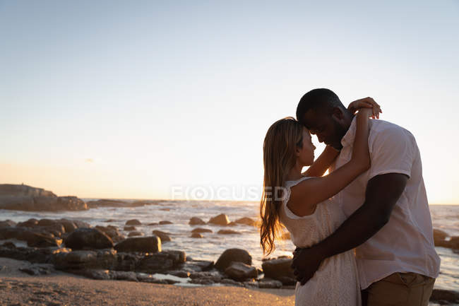 Vista lateral de casal multi étnico embaraçando uns aos outros na praia ao pôr do sol — Fotografia de Stock
