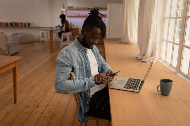 Vista lateral del hombre afroamericano usando teléfono móvil con computadora portátil en la mesa en casa - foto de stock