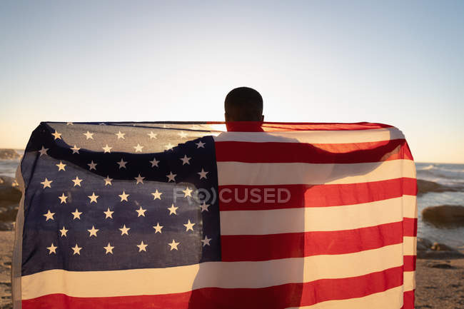 Rear view of African-American man standing at beach while holding american flag — Stock Photo