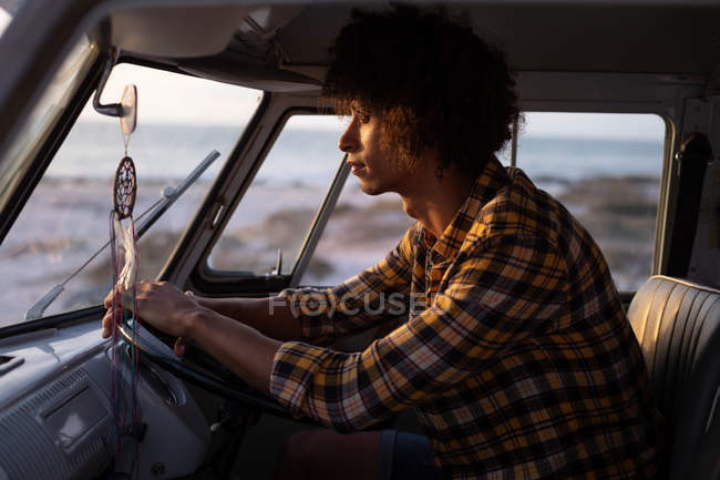 Vista lateral de un hombre de raza mixta conduciendo una caravana contra el océano en el fondo mientras se pone el sol - foto de stock