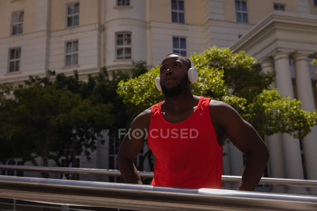 Vista frontal del reflexivo joven afroamericano en forma de hombre con auriculares de pie contra barandilla en un día soleado - foto de stock
