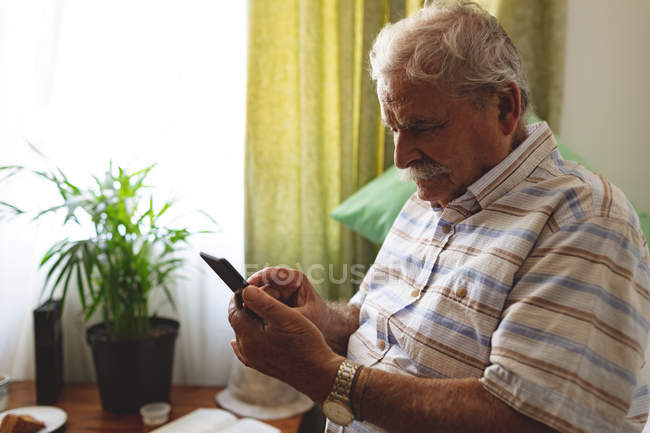Vista lateral do homem caucasiano sênior usando telefone celular enquanto sentado na cama em casa de repouso — Fotografia de Stock