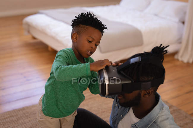 Высокоугольный вид афроамериканского сына, прикрепляющего гарнитуру виртуальной реальности к отцу дома — стоковое фото