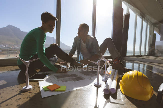 Front view of Caucasian architects sitting on office ground while they are discussing blueprints at office against a view at the office — Stock Photo