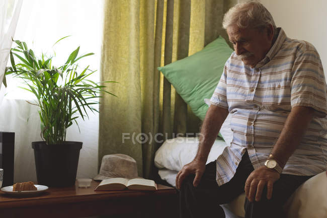 Vista frontal de un hombre mayor caucásico triste y reflexivo con la mano sobre las rodillas sentado y mirando hacia abajo en una habitación en el hogar de ancianos - foto de stock