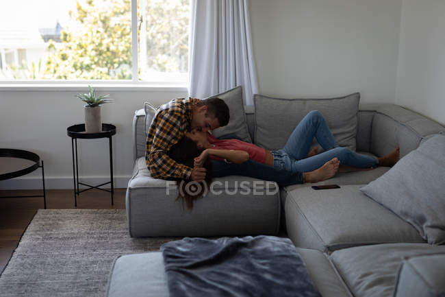 Vue latérale d'un jeune couple multi-ethnique s'embrassant alors qu'il était allongé sur un canapé à la maison — Photo de stock