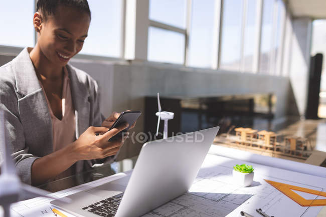 Фронтальный вид бизнесвумен смешанной расы, разговаривающей по мобильному телефону во время работы на ноутбуке за столом в офисе — стоковое фото