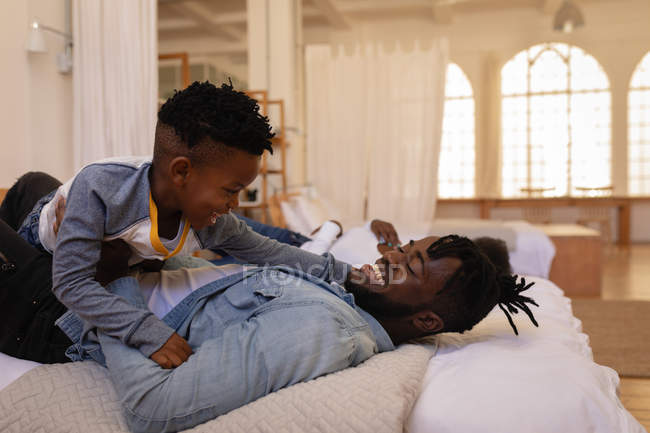 Vista lateral do feliz pai e filho afro-americano desfrutando enquanto deitado na cama em casa — Fotografia de Stock