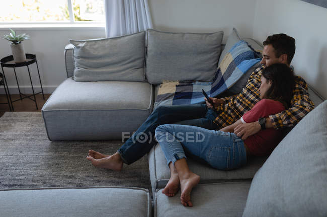 Vista lateral de la joven pareja multiétnica utilizando el teléfono móvil mientras se apoya en el sofá en casa - foto de stock