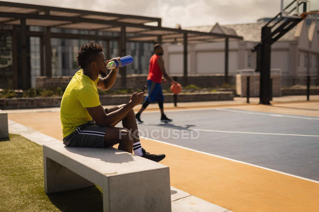 Side view of African-American basketball player drinking water while relaxing at basketball court on a stone bench against another player standing in background — Stock Photo