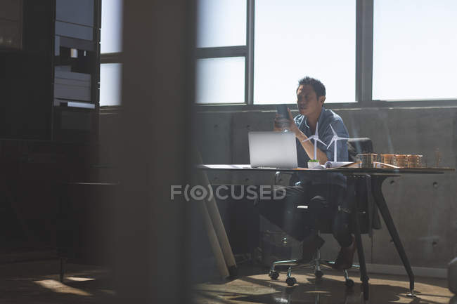 Вигляд спереду чоловічого азіатських архітектора за допомогою мобільного телефону. Сидячи на стільці в бюро в сучасні офісні — стокове фото
