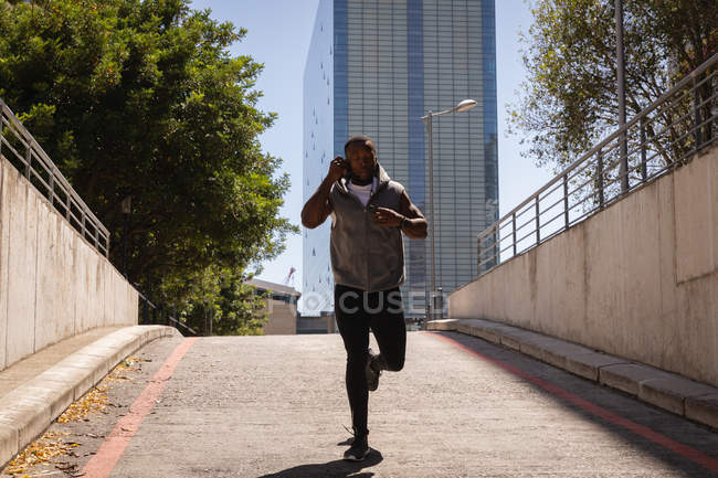 Front view of young African-American fit man talking on mobile phone while running from street on a sunny day — Stock Photo
