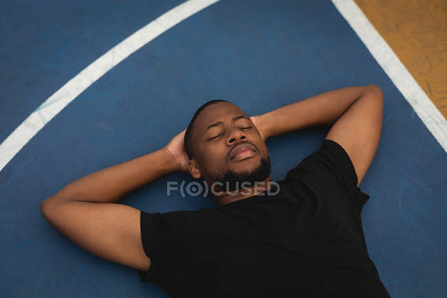 Vista de ángulo alto del joven jugador de baloncesto afroamericano con las manos detrás de la cabeza y los ojos cerrados relajándose en el suelo en la cancha de baloncesto - foto de stock
