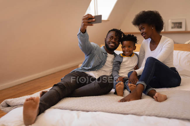 Vista frontal de la feliz familia afroamericana sentados juntos y tomando selfie en casa. Están sonriendo y mirando el teléfono móvil - foto de stock