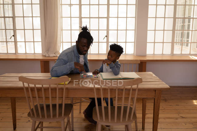 Frontansicht eines afrikanisch-amerikanischen Vaters, der seinem Sohn beim Zeichnen hilft, während er im Esszimmer auf einem Stuhl sitzt — Stockfoto