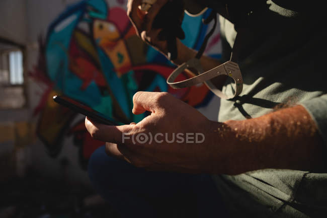 Sección media del joven artista de graffiti caucásico usando el móvil mientras está sentado en el callejón - foto de stock