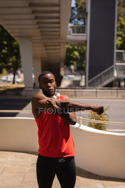 Вид спереди на молодого афроамериканца, занимающегося растяжкой рук на тротуаре под мостом в солнечный день — стоковое фото