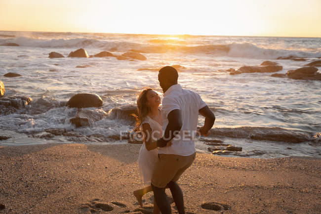 Vista laterale di felice coppia multietnica godendo e giocando sulla spiaggia al tramonto — Foto stock