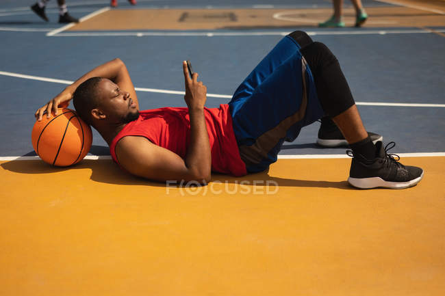 Vista lateral do jogador de basquete afro-americano deitado no chão do parque infantil com a cabeça apoiada no basquete enquanto ele está usando o telefone celular na quadra de basquete — Fotografia de Stock