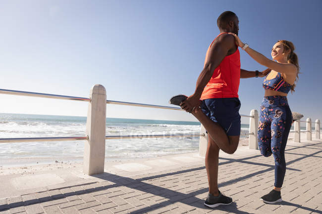 Vue de côté du jeune couple multi-ethnique faisant de l'exercice sur la chaussée près de la plage par une journée ensoleillée — Photo de stock