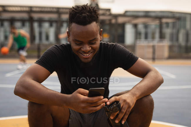 Vista frontal do jogador de basquete afro-americano sentado em um basquete enquanto ele está usando o telefone celular em sorrir em um campo de basquete contra jogadores em segundo plano — Fotografia de Stock