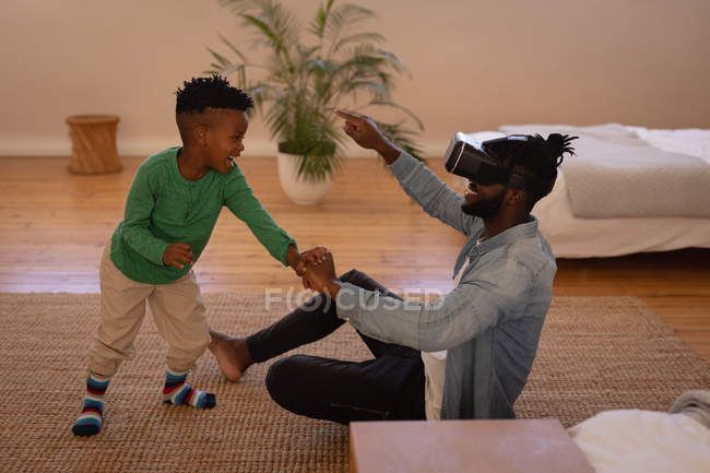Vue latérale du père et du fils afro-américains heureux tout en utilisant un casque de réalité virtuelle à la maison — Photo de stock