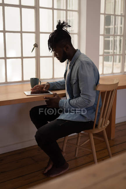 Vista lateral del hombre afroamericano usando tableta digital mientras está sentado en la silla en casa - foto de stock