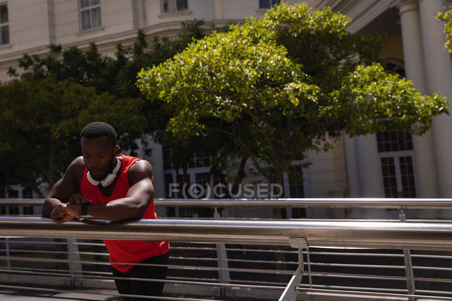 Vista frontale del giovane afro-americano in forma uomo con auricolare guardando orologio intelligente mentre in piedi contro ringhiera — Foto stock