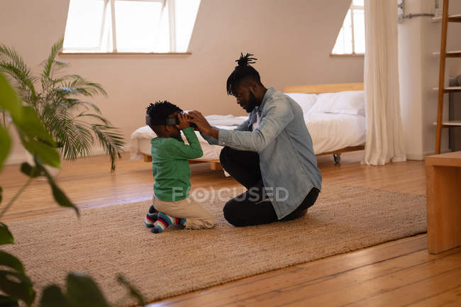 Вид сбоку красивого афро-американского отца, помогающего сыну использовать гарнитуру виртуальной реальности дома — стоковое фото