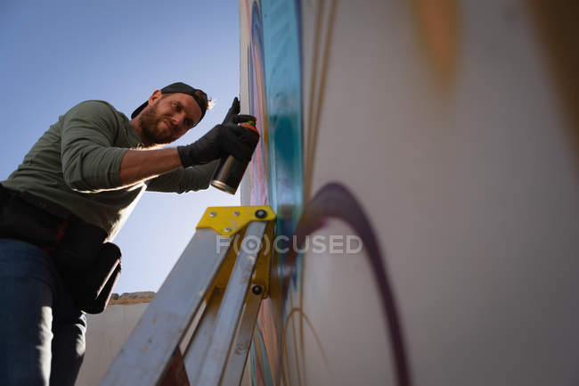 Vue à angle bas de jeune artiste graffiti caucasien peinture par pulvérisation sur chambre murale altérée — Photo de stock