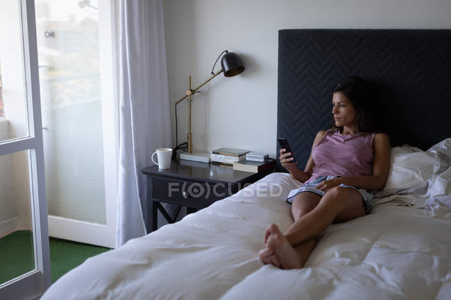 Вид спереду красива жінка змішаної раси, що використовує мобільний телефон, лежачи на ліжку вдома — стокове фото