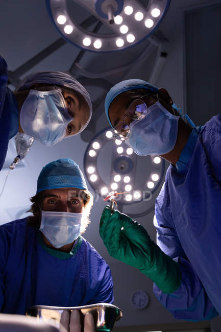 Low angle view of multi-ethnic surgeons concentrated performing operation in operating room at hospital with lights at ceiling — Stock Photo