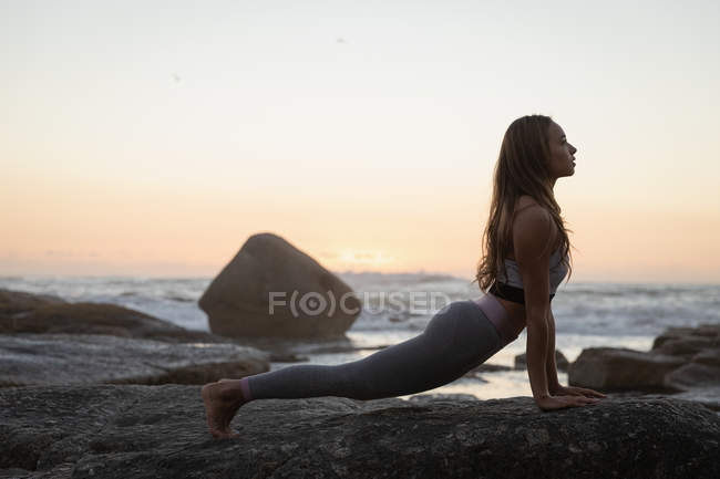 Vista lateral de la mujer haciendo yoga sobre roca en la playa al atardecer - foto de stock